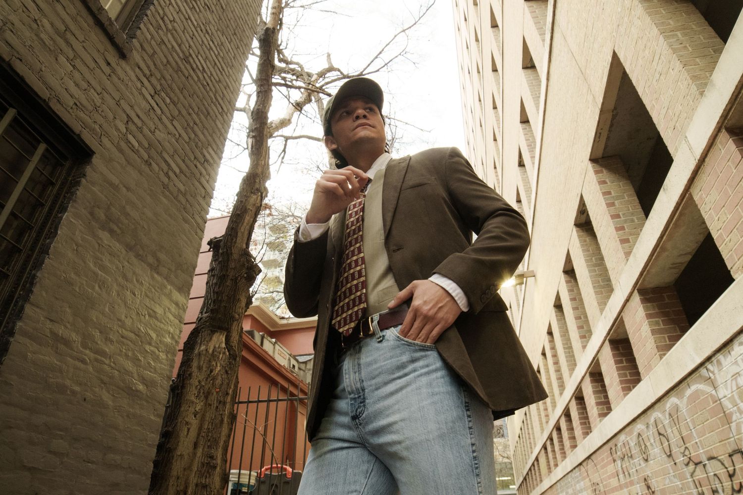 Low angle shot of man in brown blazer and cap in urban alleyway setting.