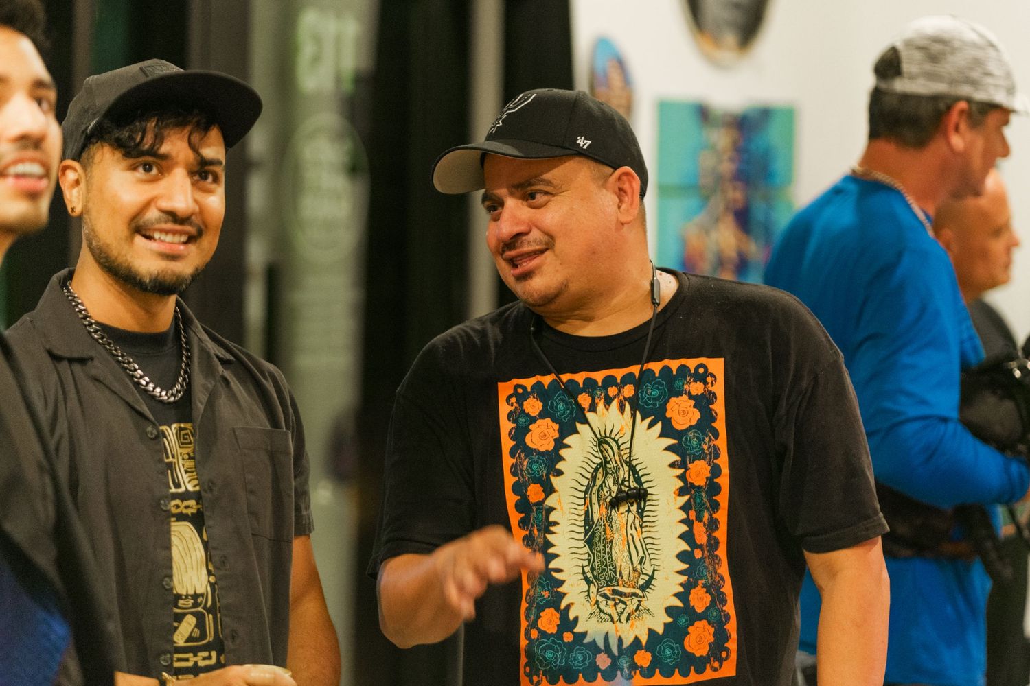 Two men smiling and conversing at an indoor art event, one wearing a Guadalupe graphic t-shirt.
