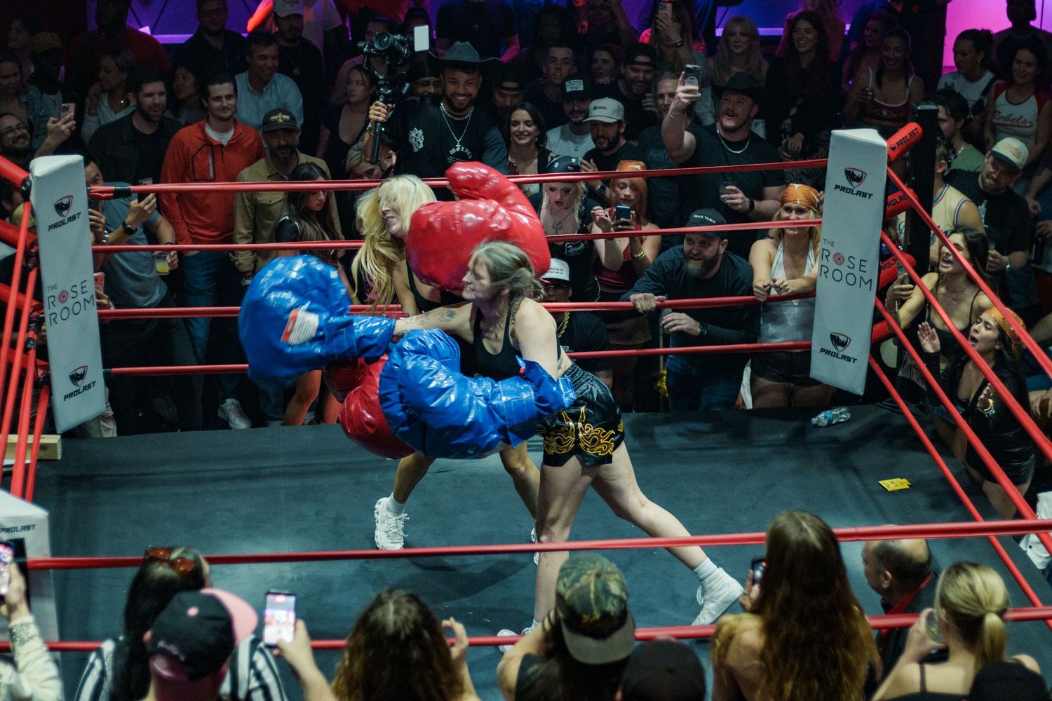 Two women in oversized boxing gloves exchange blows in a boxing ring surrounded by an enthusiastic crowd.