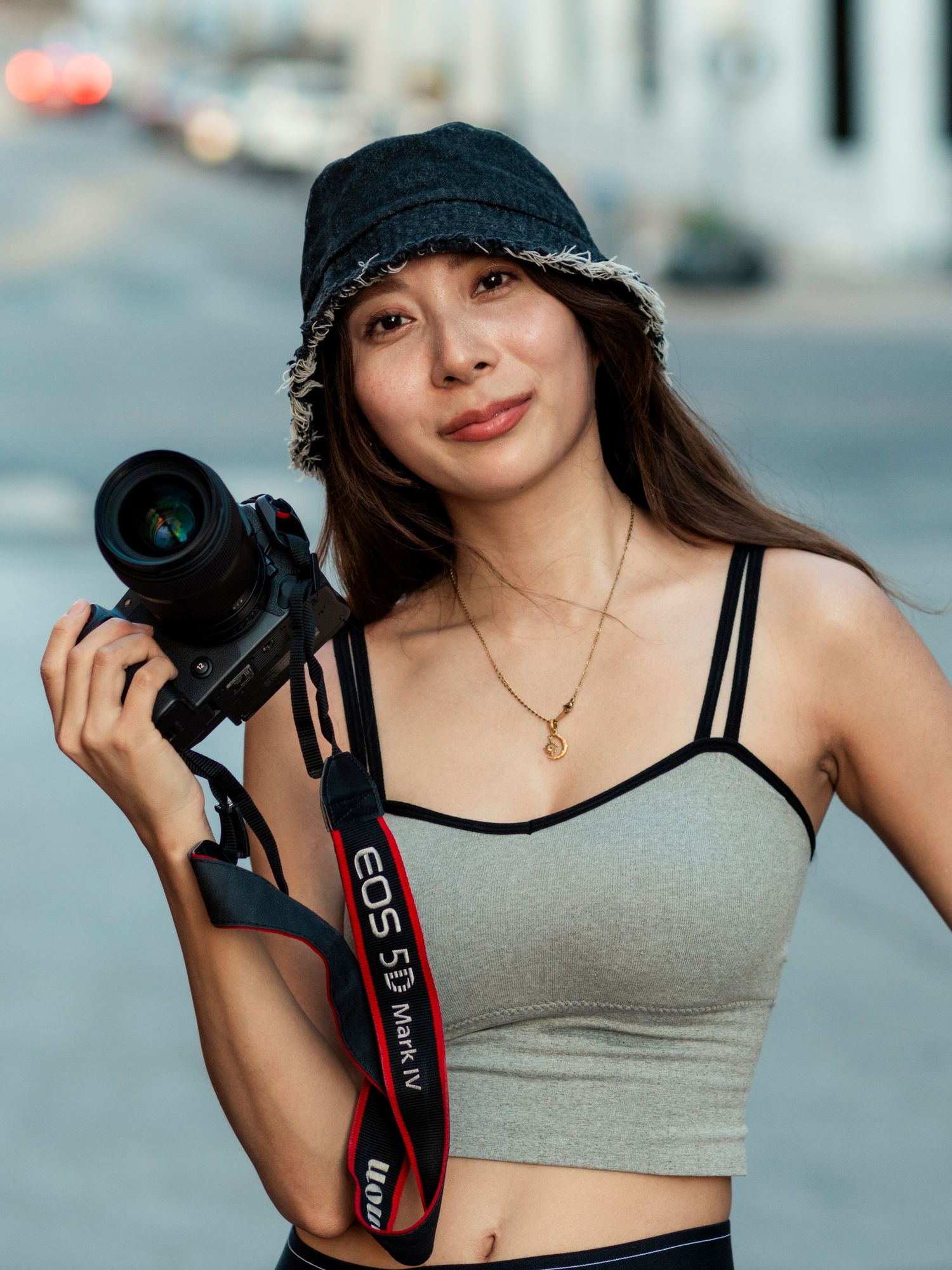 Young woman in bucket hat holding Canon EOS 5D Mark IV camera on city street.