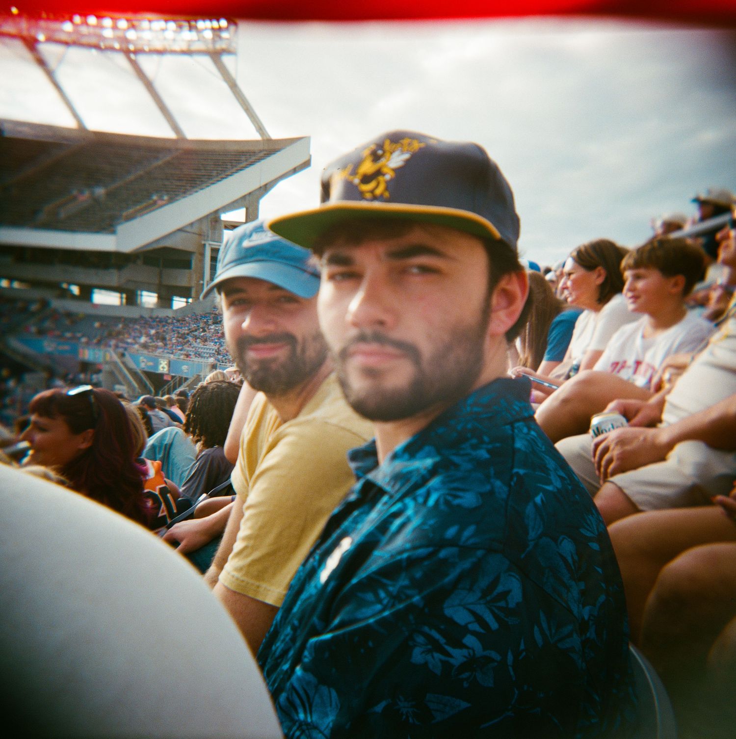 Two bearded men wearing baseball caps sit in a packed outdoor stadium on a sunny day.