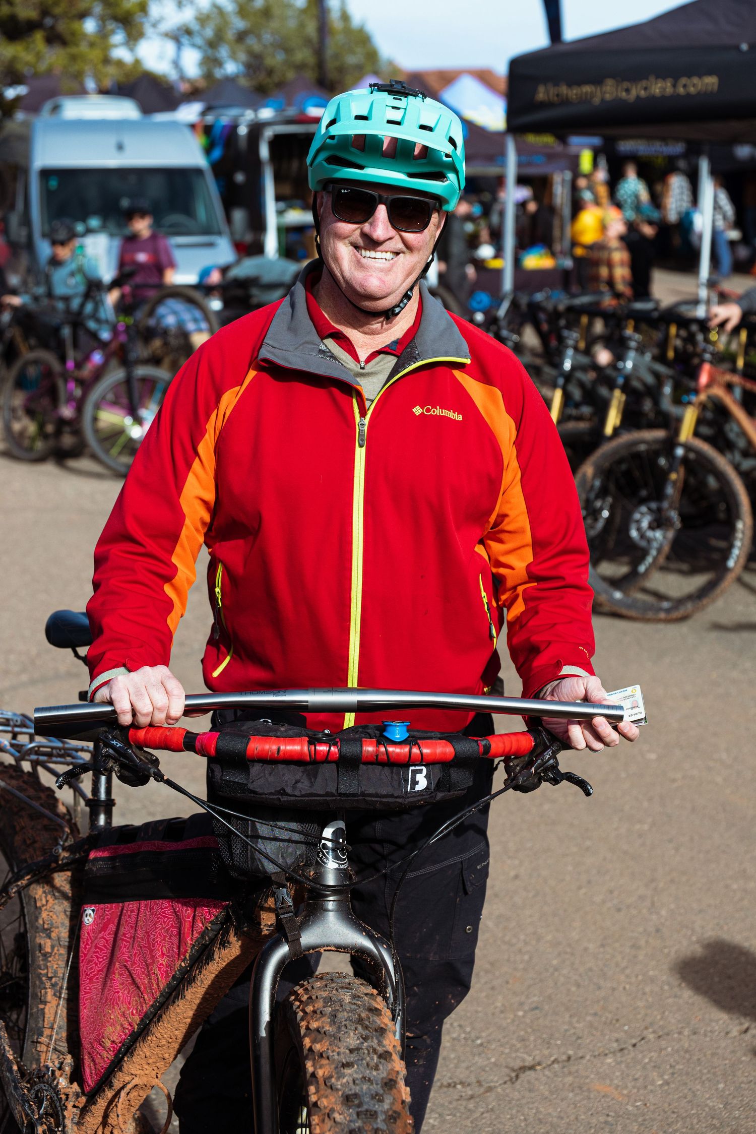 Happy cyclist in red jacket and teal helmet posing with a mountain bike at an outdoor bike expo.