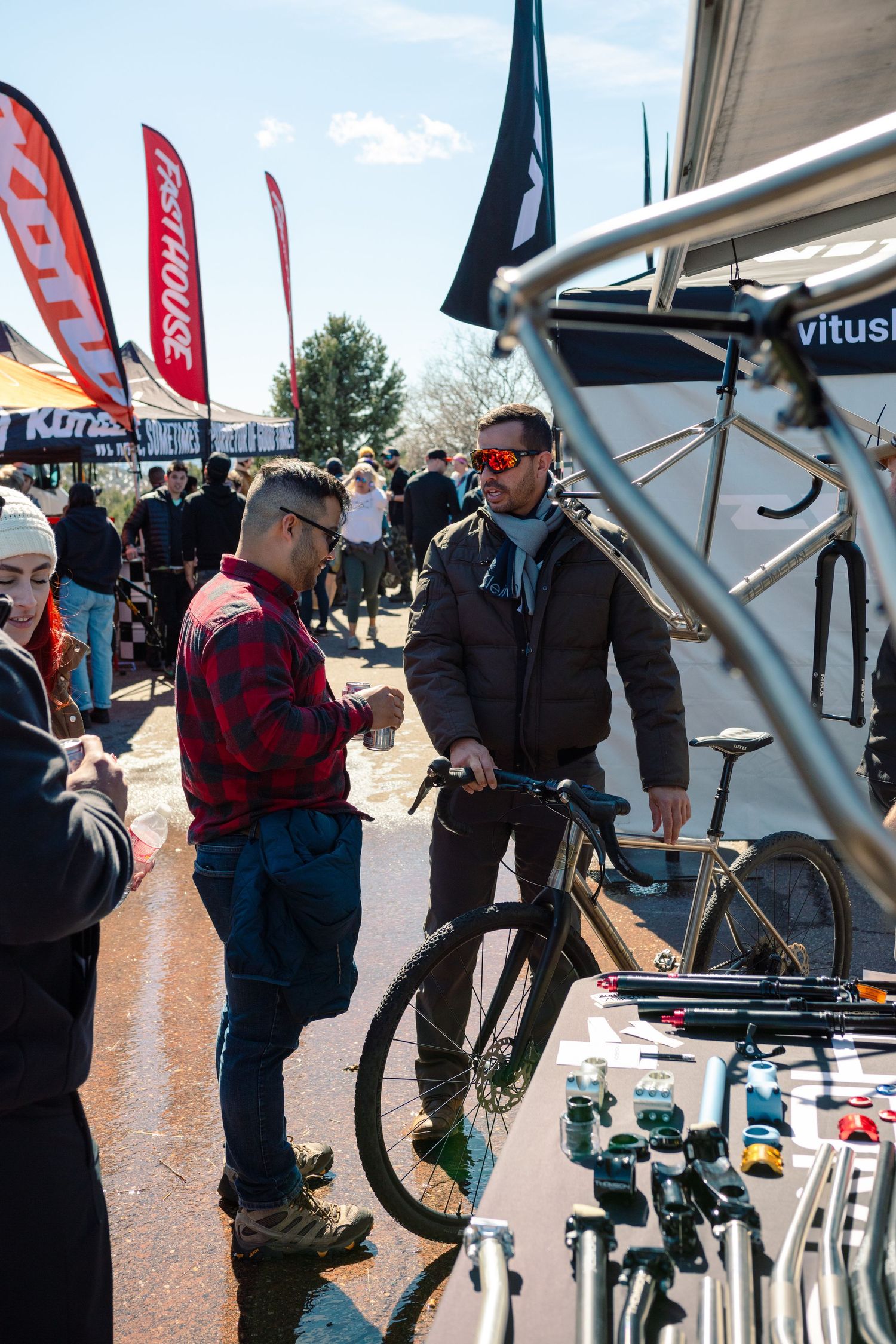 Two men discuss a bicycle at an outdoor cycling trade show near a Vitus Bikes booth.