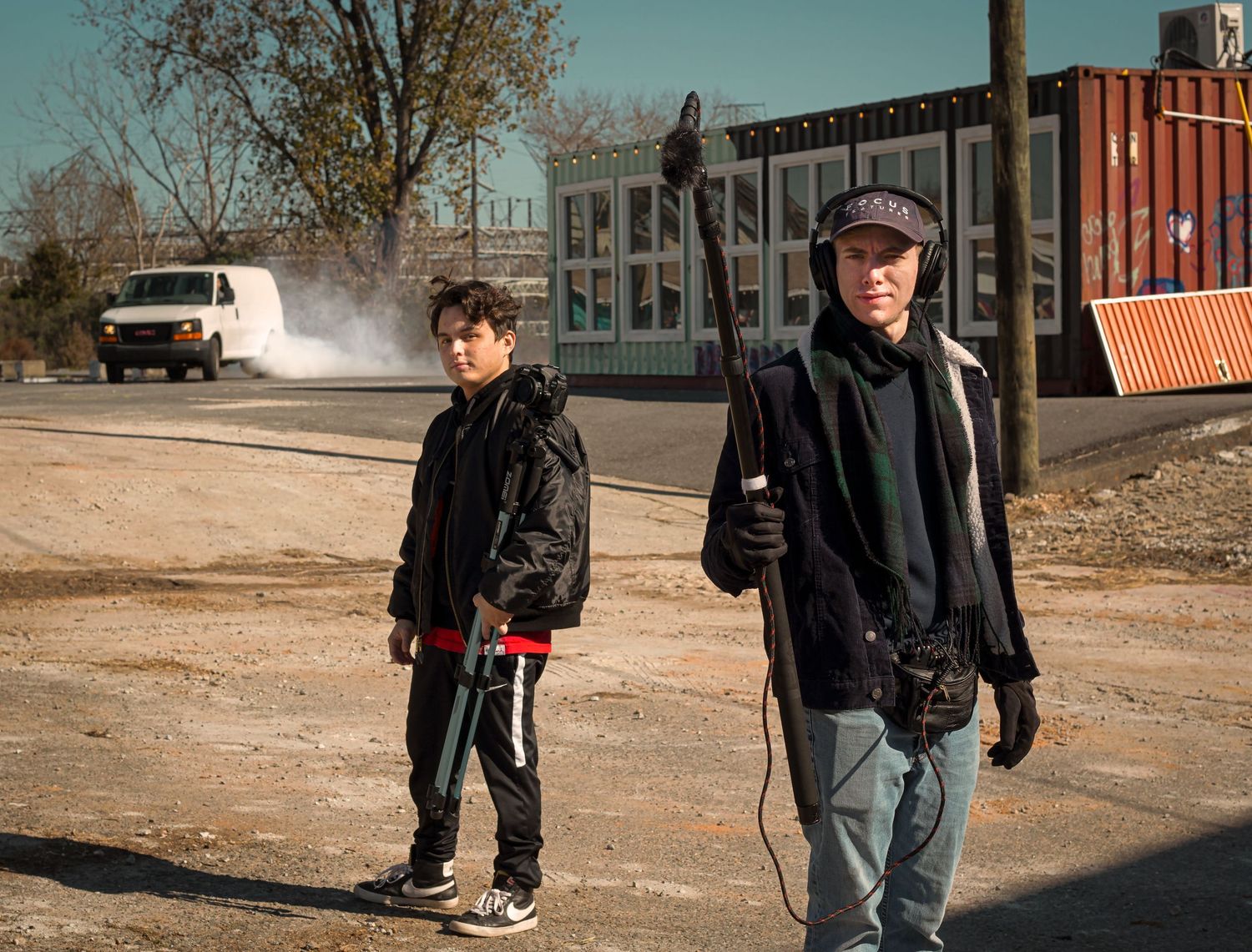 Two men standing outdoors in an industrial area, one holding a tool, with a white van and shipping containers in the background.