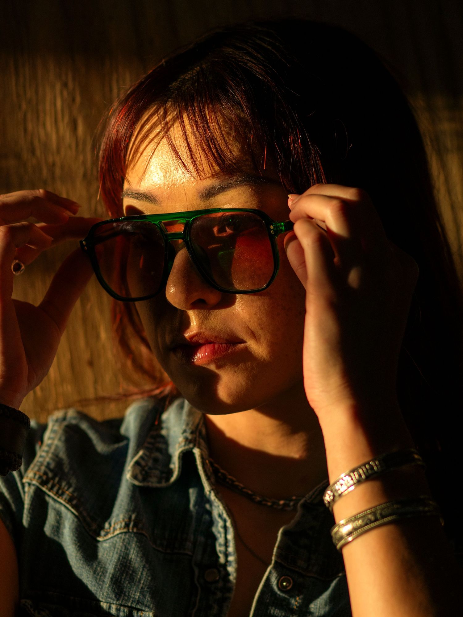 Young Asian woman with red hair adjusting green-framed sunglasses in dramatic warm lighting.