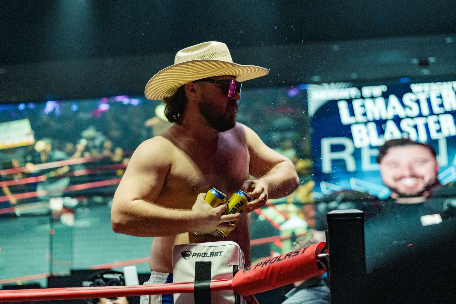 Shirtless male boxer wearing cowboy hat and sunglasses stands confidently in a boxing ring corner.