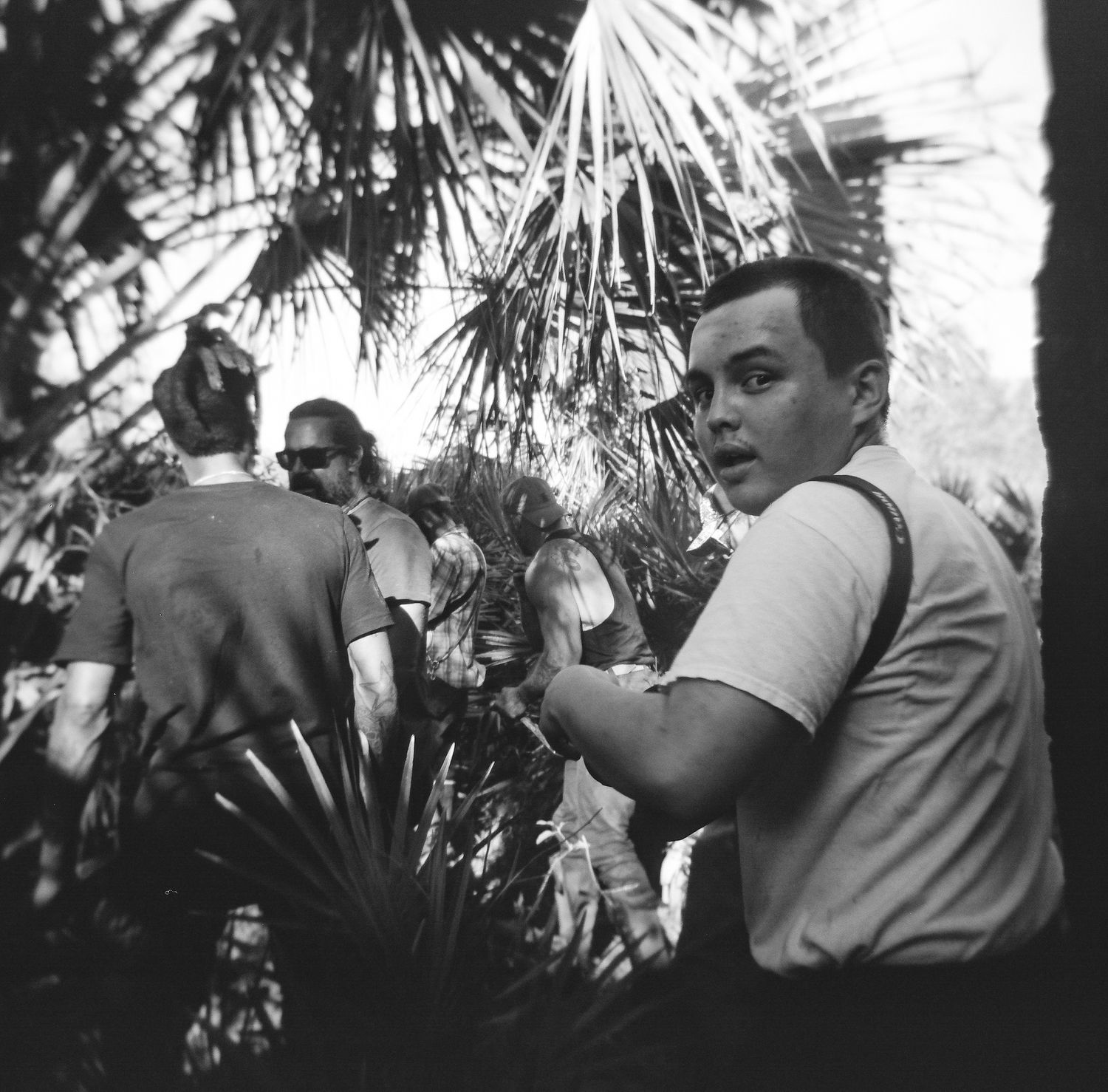 Black and white photo of a young man looking back at camera surrounded by tropical palm plants.