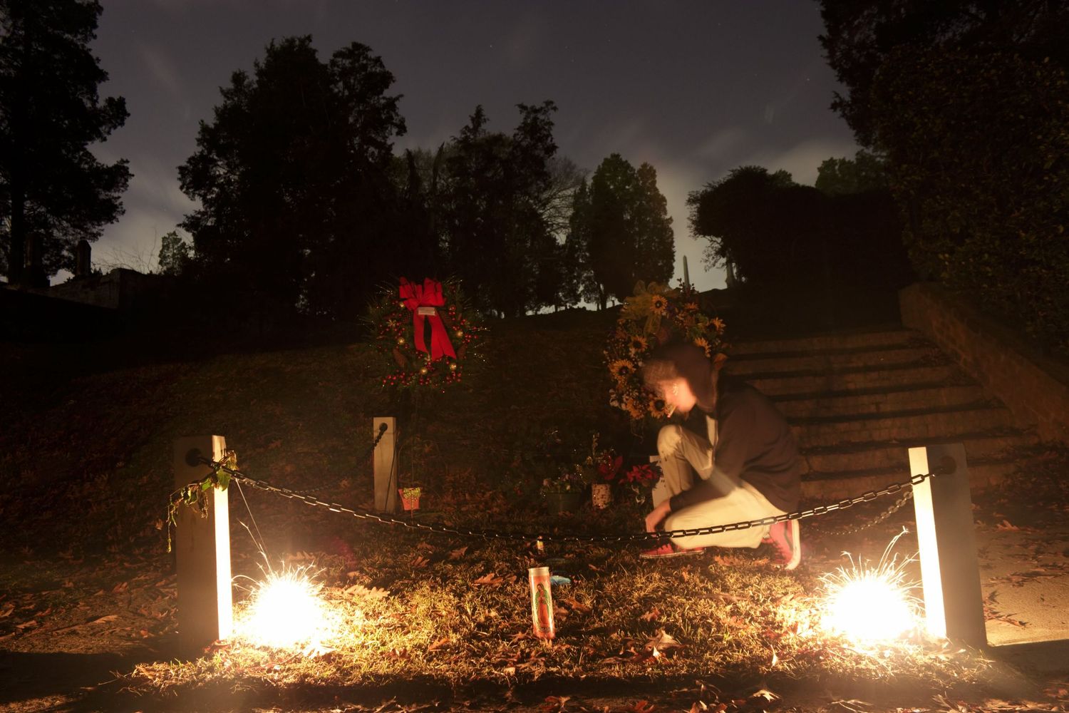 Person kneeling at an outdoor memorial at night, illuminated by candles and sparkling lights among autumn leaves.