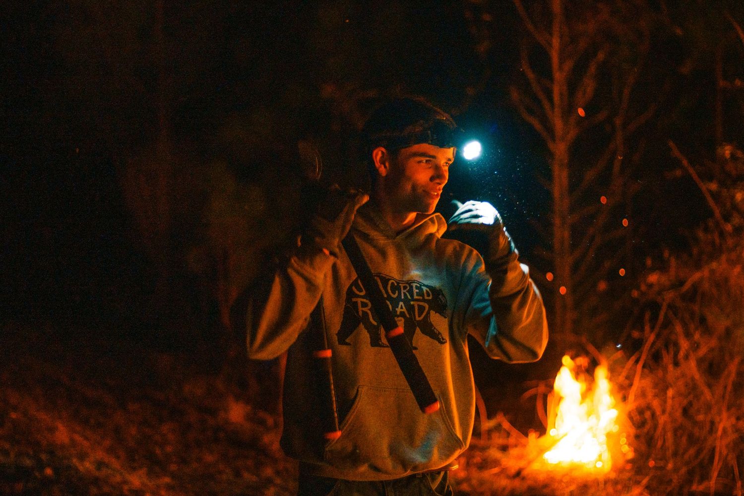 Man in hoodie holding flashlight stands by campfire at night in dark forest wearing headlamp outdoors.