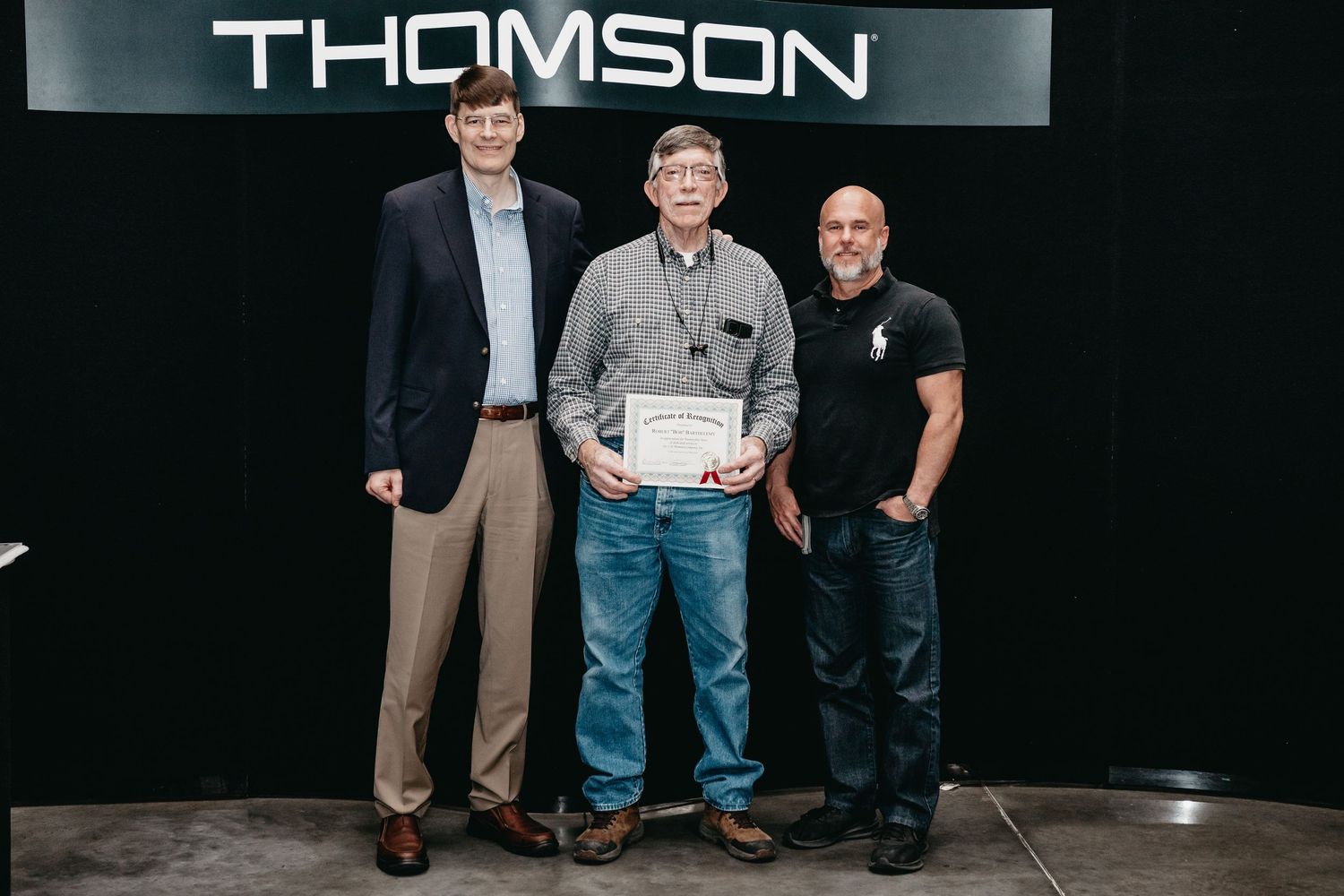 Three men pose for a photo in front of a Thomson sign, with the middle man holding a certificate award.