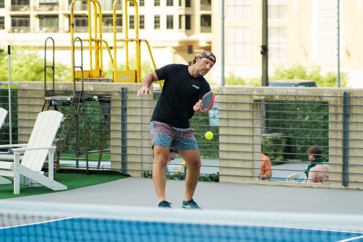 Man playing pickleball on an outdoor court, mid-swing hitting a yellow ball wearing black shirt and patterned shorts.