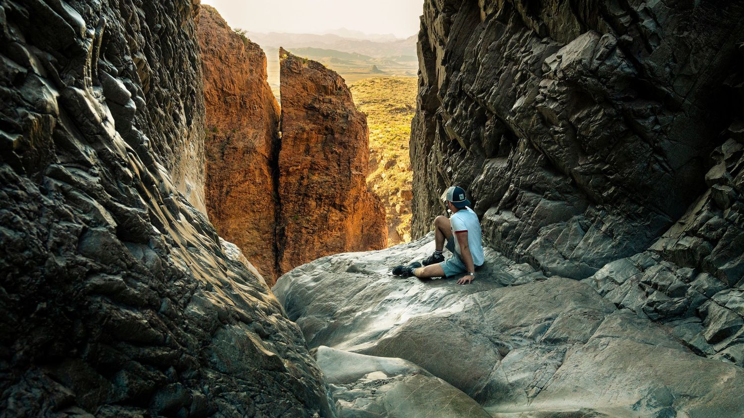 Solo traveler resting in a scenic rocky canyon slot with vast arid desert terrain in the background.