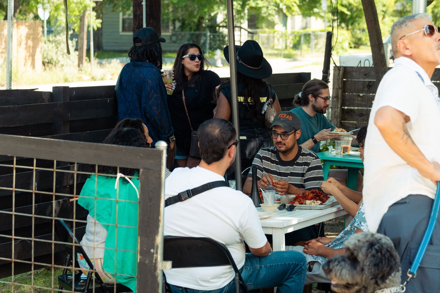 People eating and socializing at outdoor tables under trees at a casual daytime gathering.
