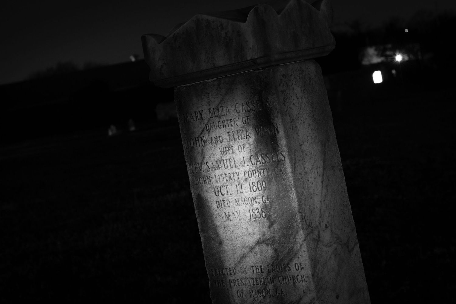 Black and white nighttime photo of an old weathered gravestone in a dark cemetery.