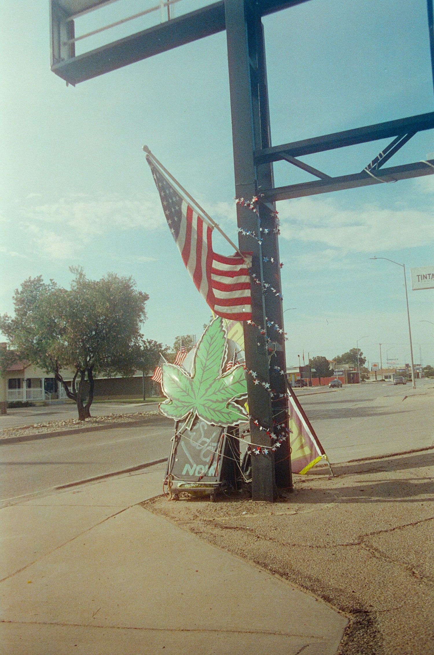 American flag and cannabis leaf sign displayed on a decorated pole at a street corner on a sunny day.