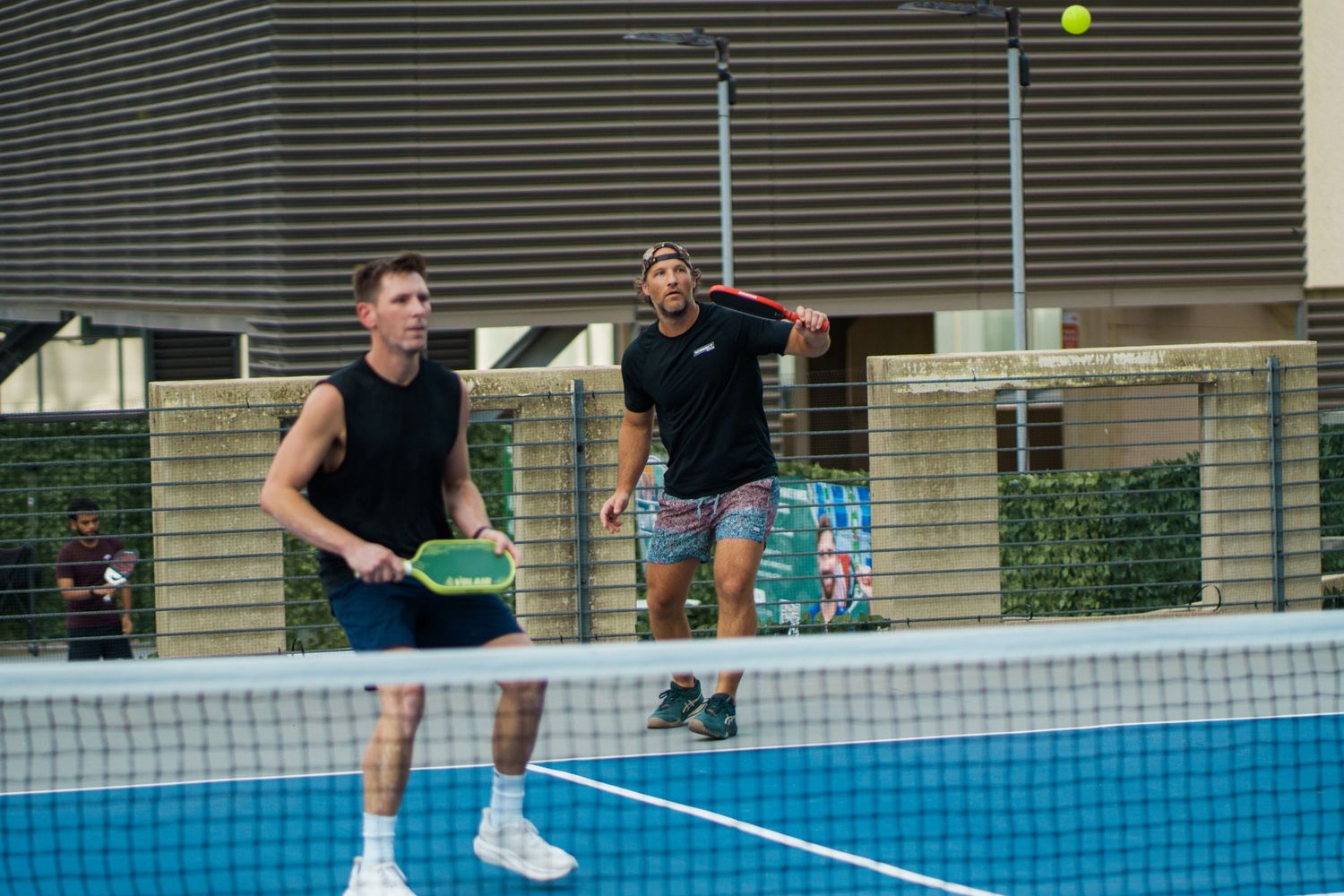 Two men playing doubles padel tennis on an outdoor court, one swinging at a yellow ball mid-air.