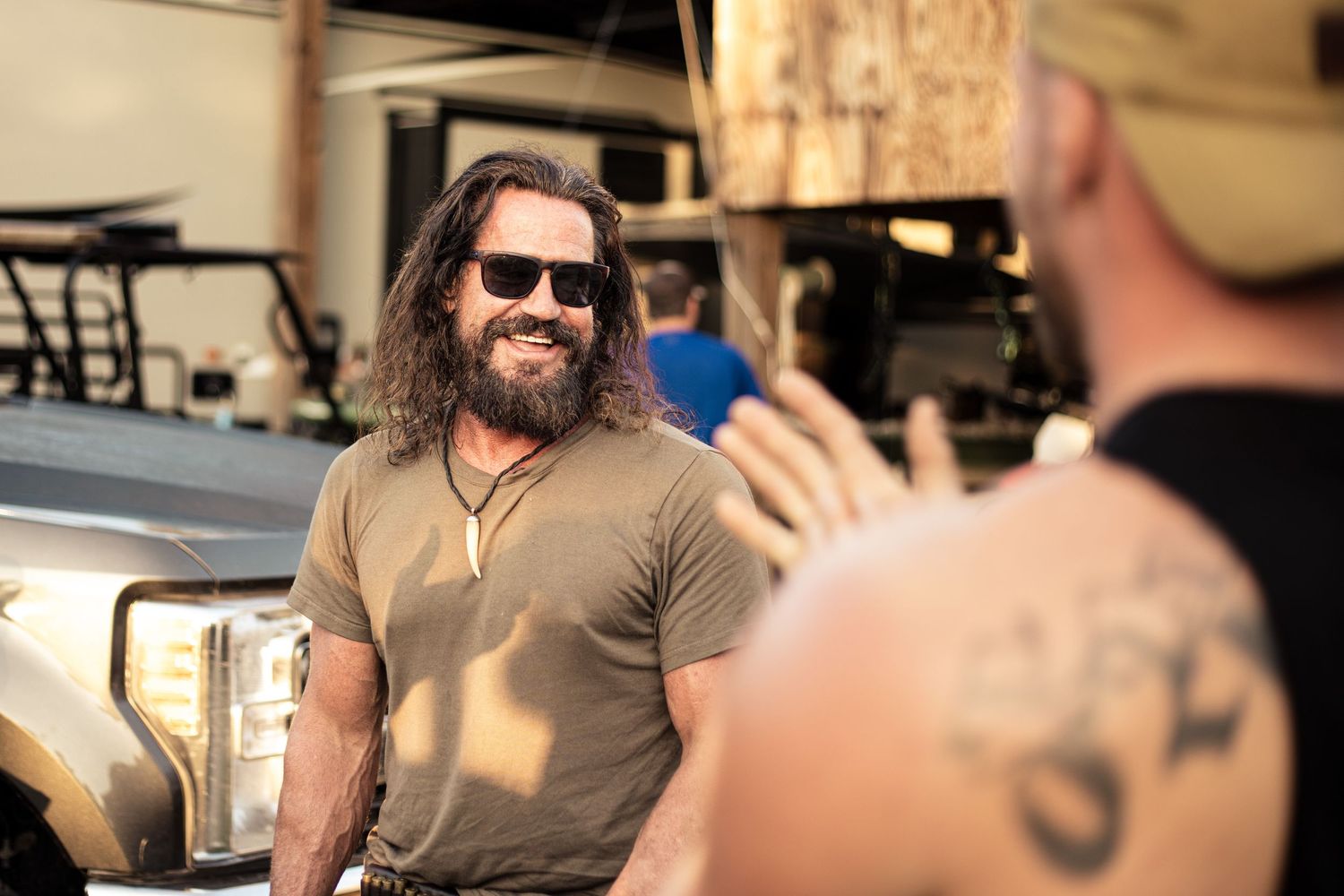 Bearded man with sunglasses smiling outdoors near a vehicle, talking to a tattooed person in natural light.