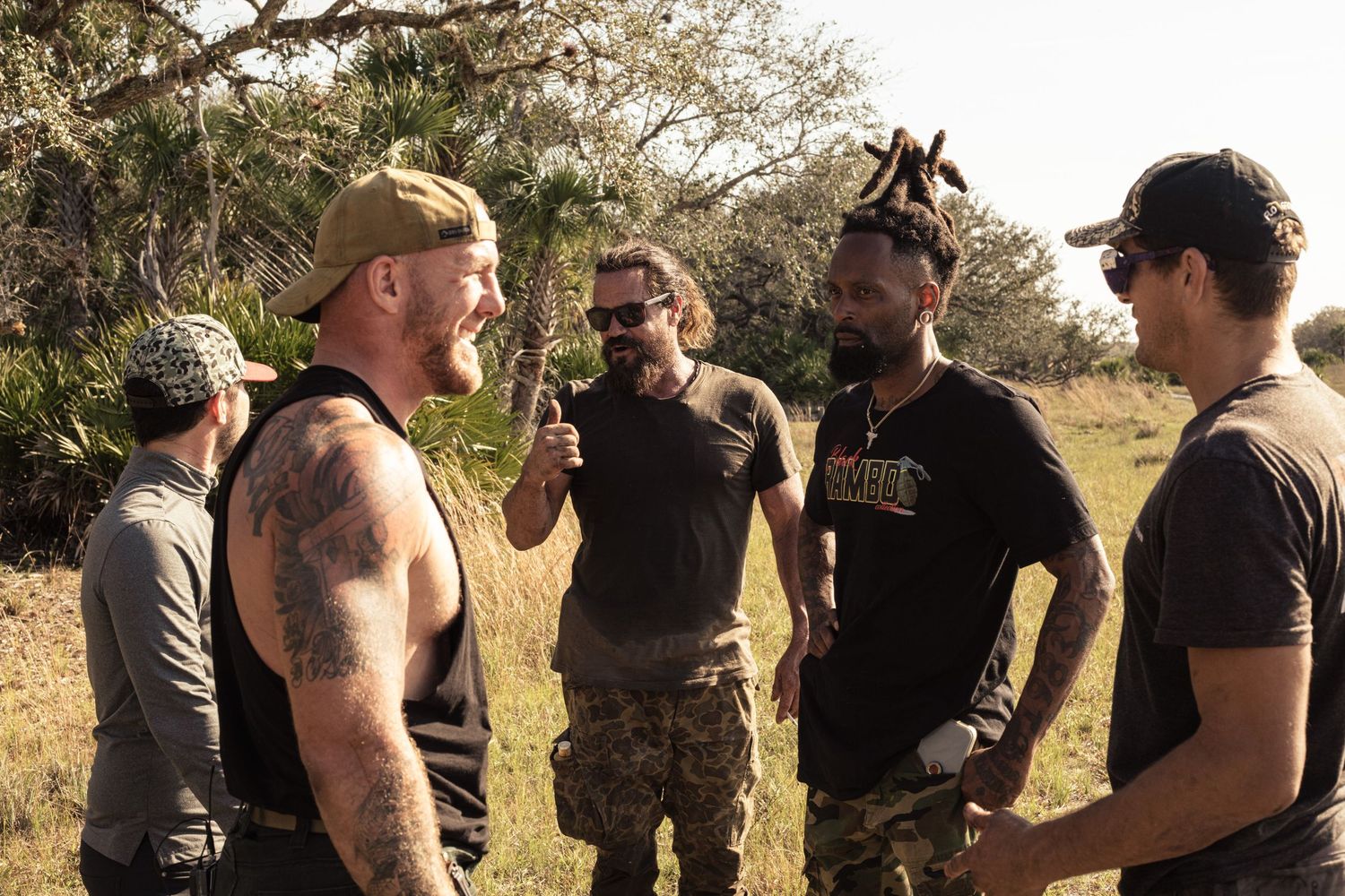Group of men in tactical gear conversing outdoors in a grassy field on a sunny day.