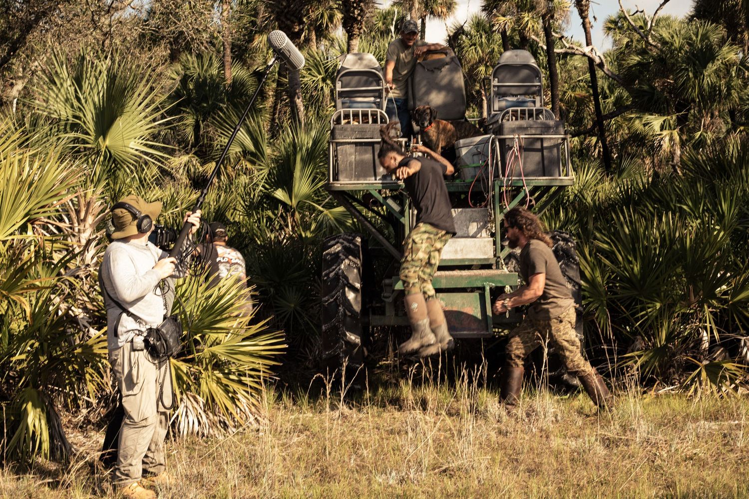 Film crew shooting a scene near a large tractor in a wild grassy landscape with palm trees.