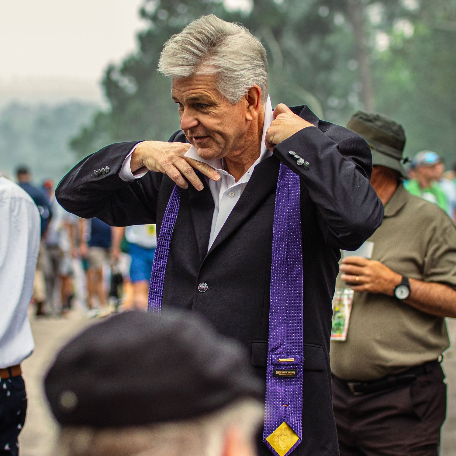 Man in black blazer adjusting purple graduation stole outdoors at a ceremony event.