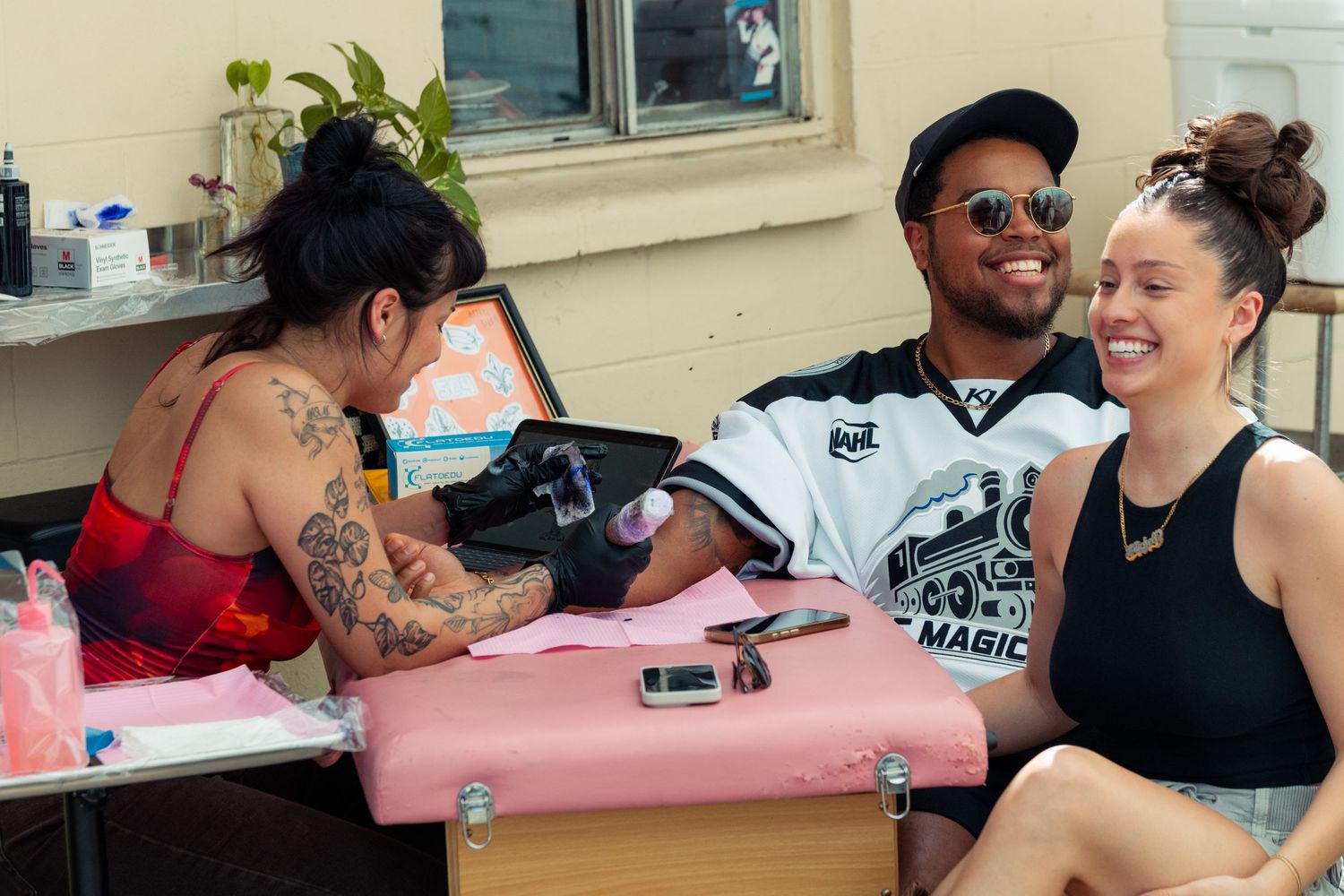 Smiling man getting a tattoo on his arm by a heavily tattooed artist while a woman sits nearby.
