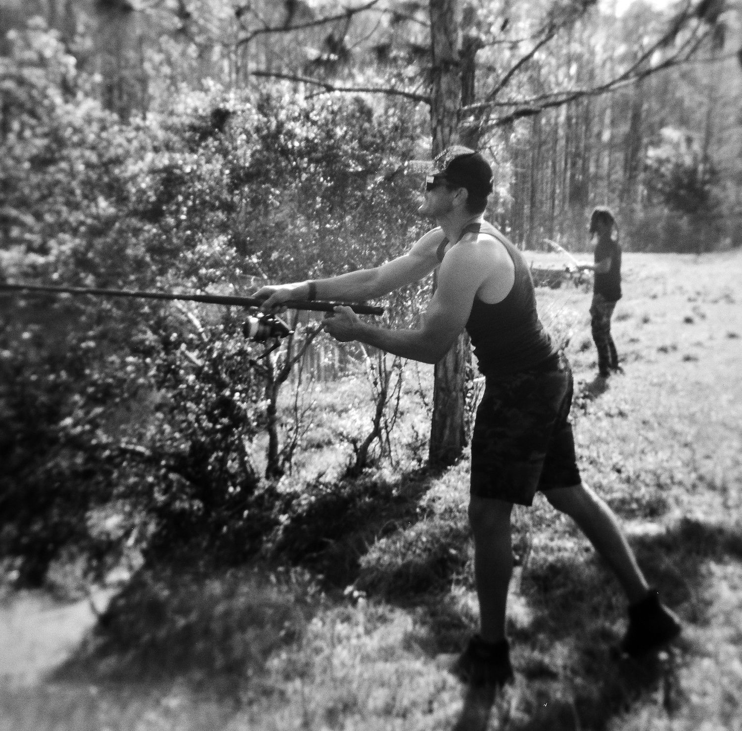 Man in tank top casts a fishing rod in a pine forest with another person in the background.