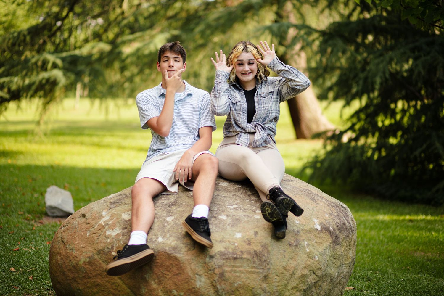 Two teenagers sitting on boulder making silly faces in outdoor park setting.
