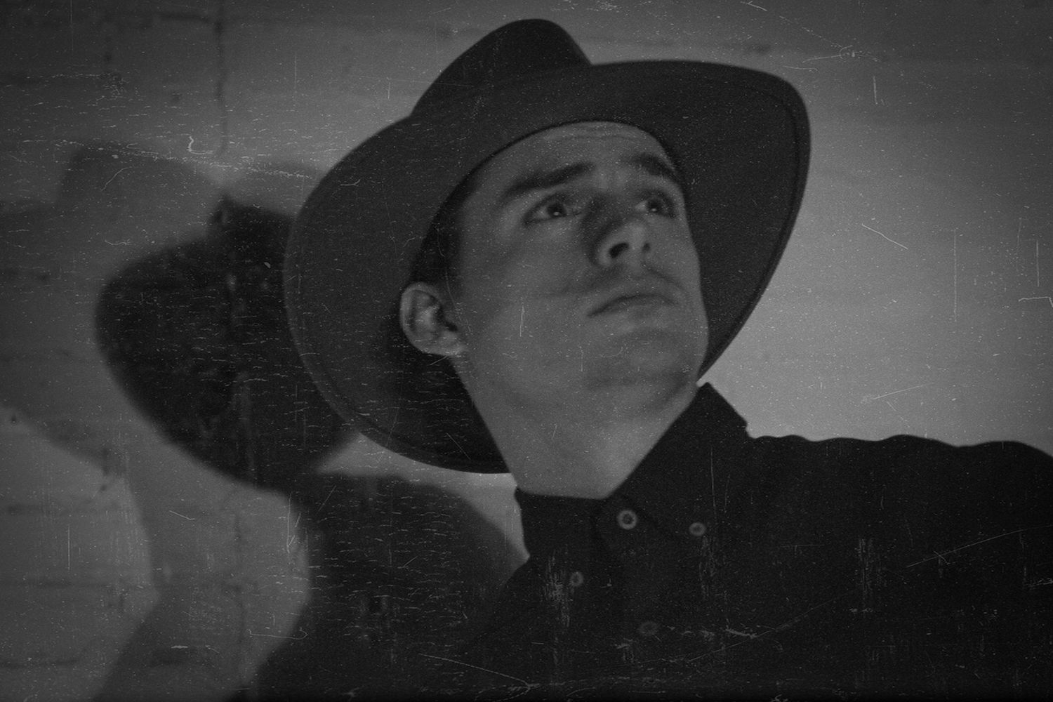 Black and white photo of a young man wearing a cowboy hat, leaning against a textured wall.
