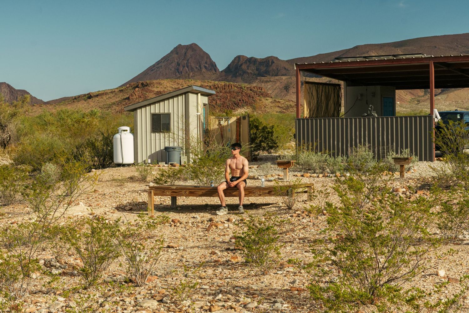 A shirtless person sits on a bench near rustic desert buildings with mountains in the background.