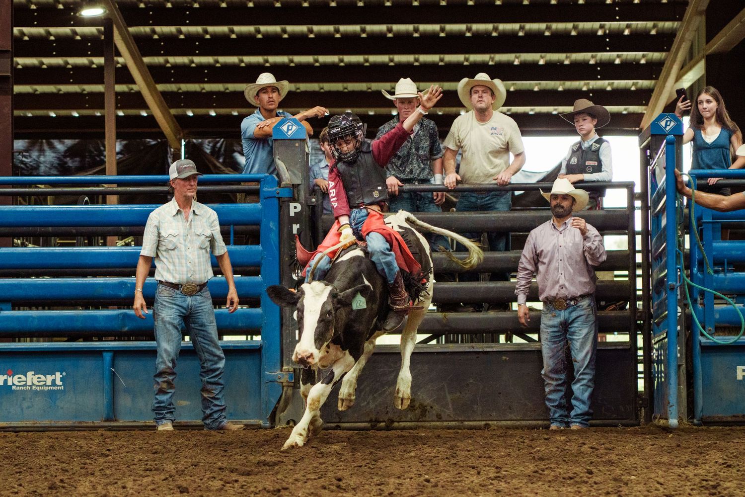 Bull rider competing at an indoor rodeo arena with cowboys and handlers watching from the fence.