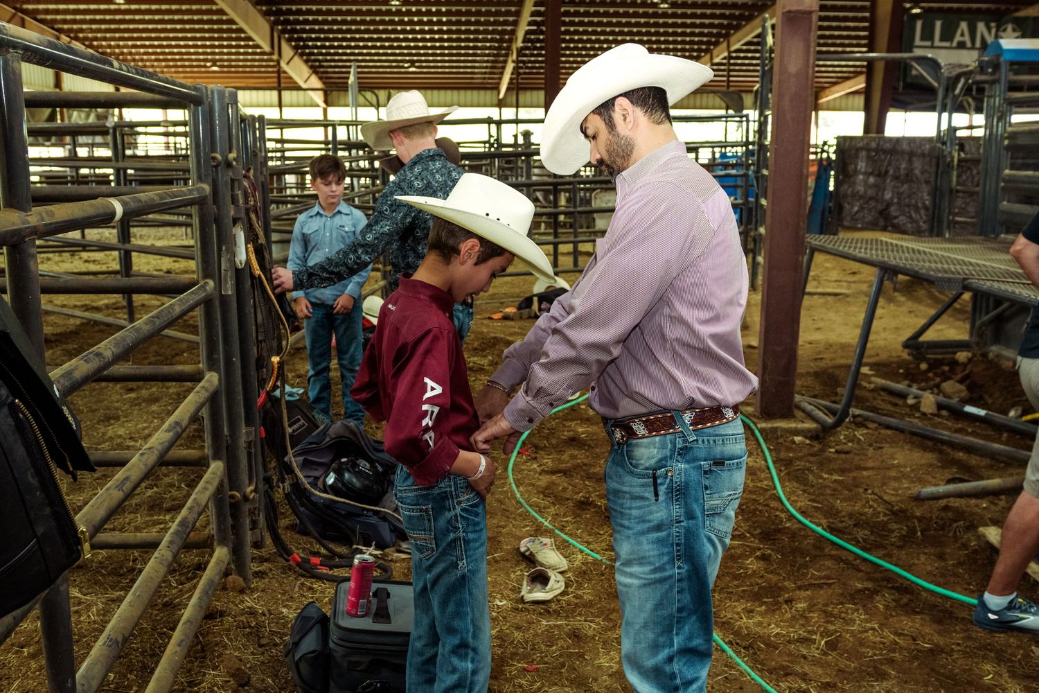 Cowboys in white hats preparing for a rodeo event inside a livestock barn with cattle pens.