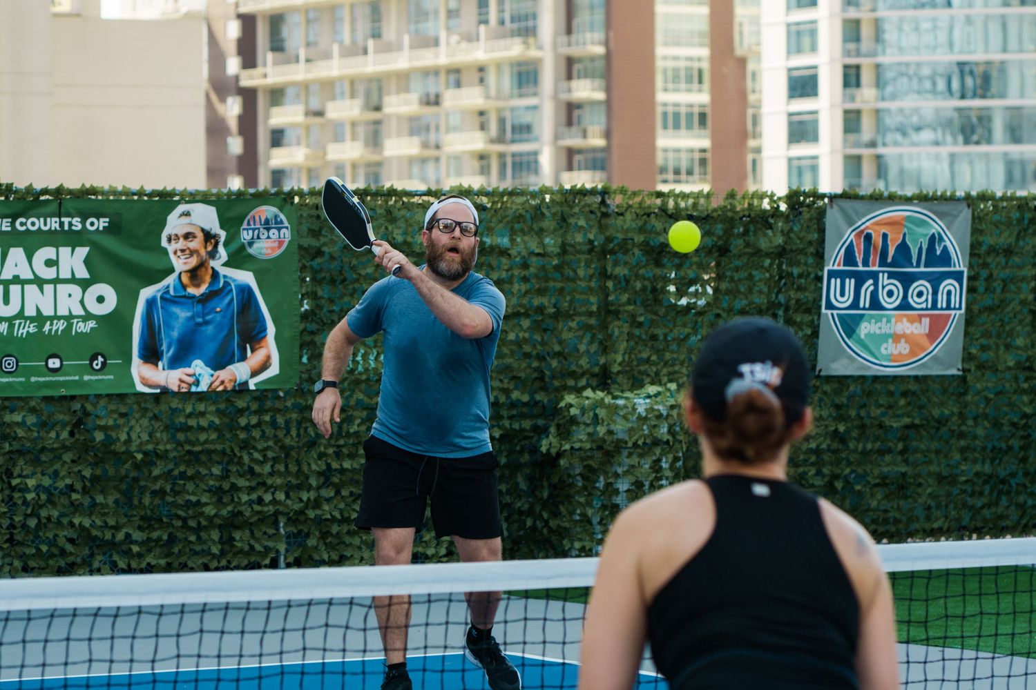 Two players enjoy a pickleball match on an outdoor urban court with a green hedge backdrop.