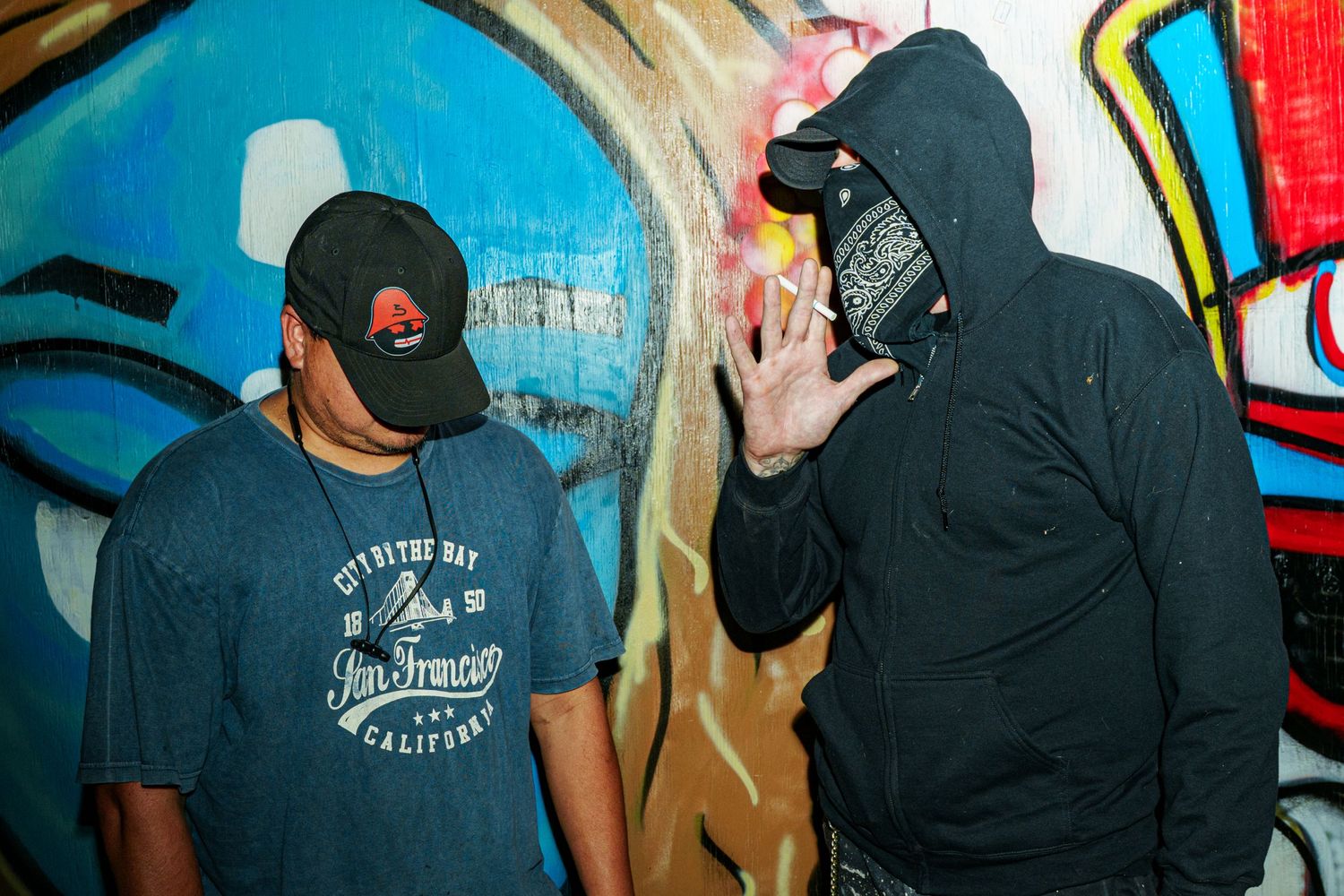 Two men in caps pose near colorful graffiti wall, one in black hoodie gesturing with hand.