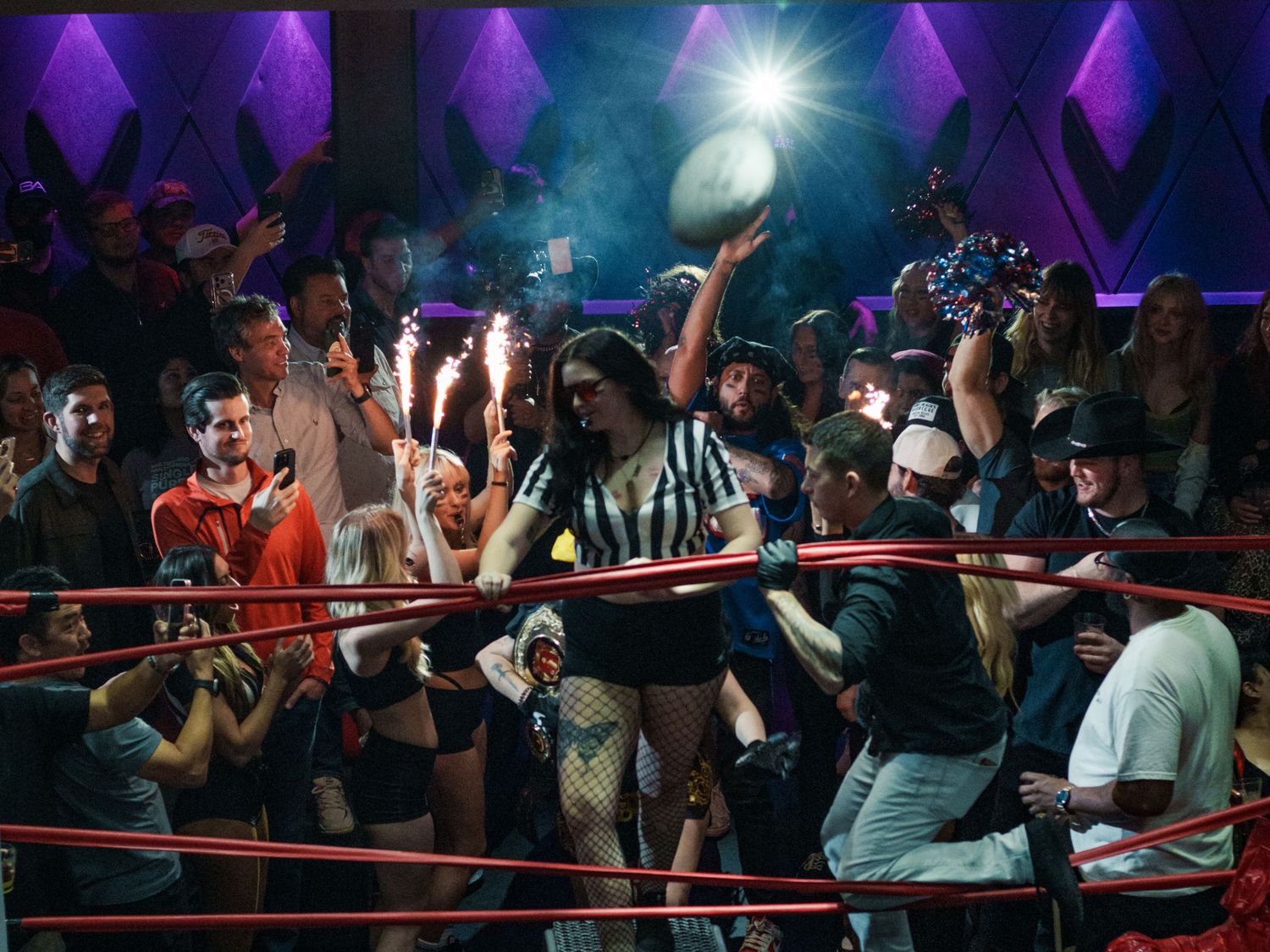 Referee tosses football in a festive wrestling ring surrounded by excited fans with sparklers.