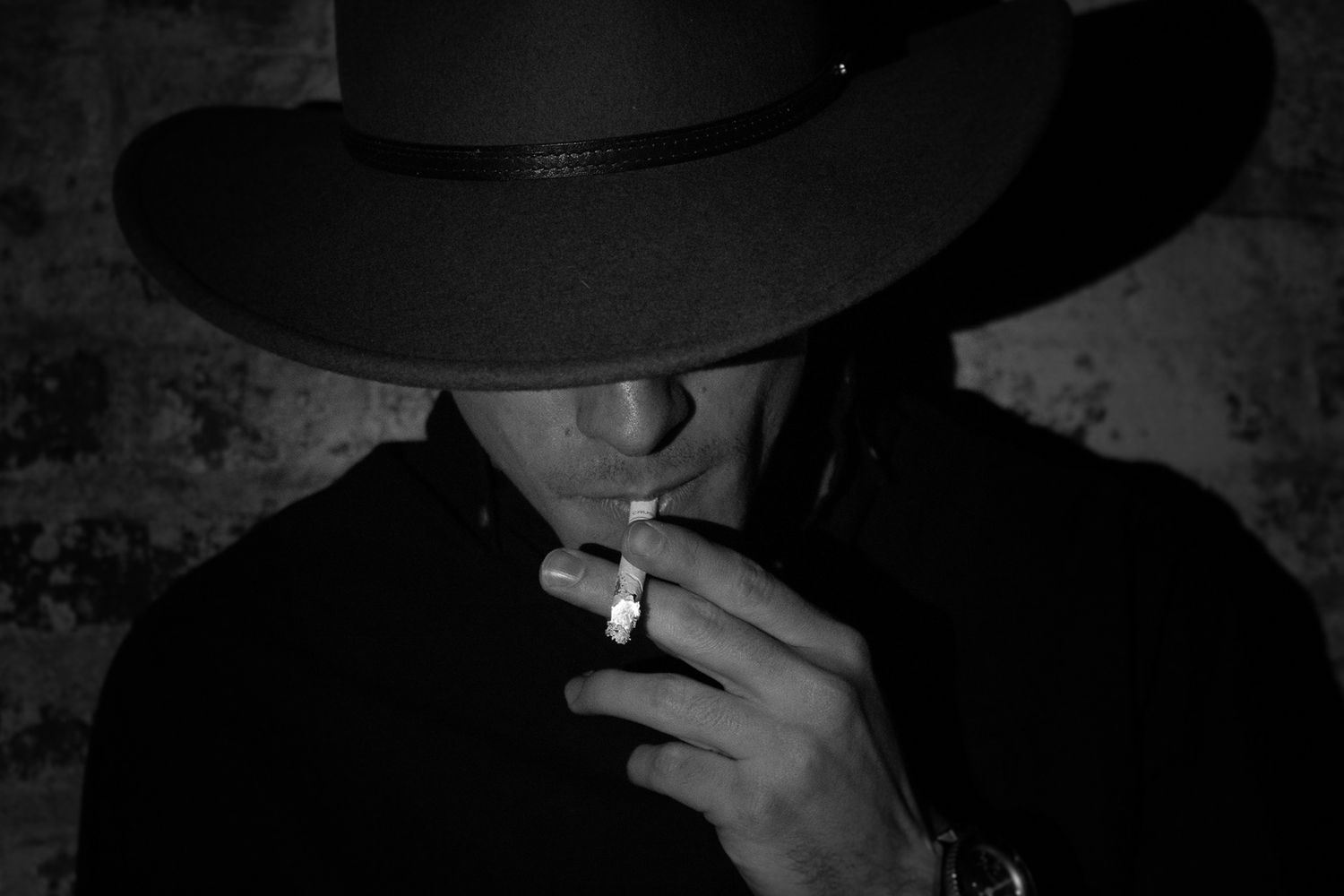 Person wearing wide brim hat smoking cigarette in moody black and white portrait.