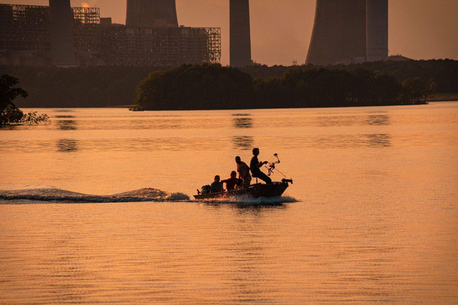 Silhouetted boat with passengers cruising on golden river at sunset with industrial cooling towers in background.