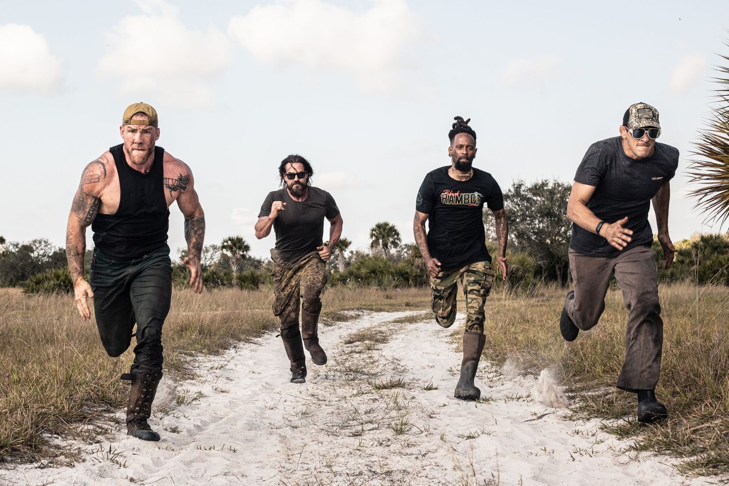 Four men running on a sandy dirt path through a grassy field on a cloudy day.