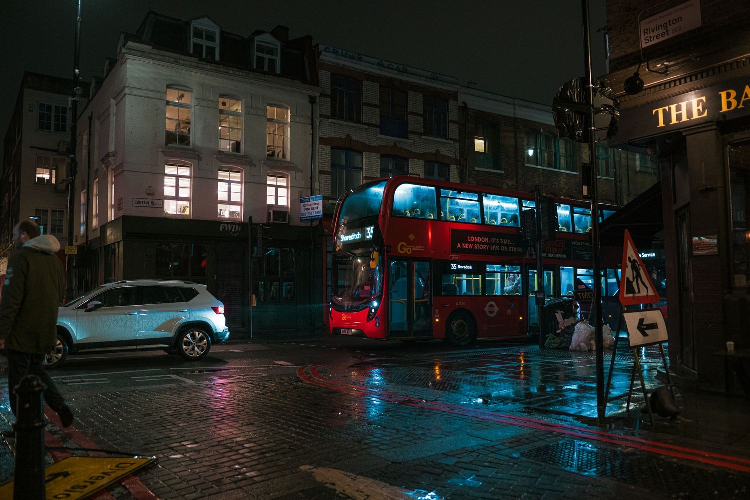 Red double-decker bus on a rainy London street at night, reflecting lights on wet pavement near historic buildings.