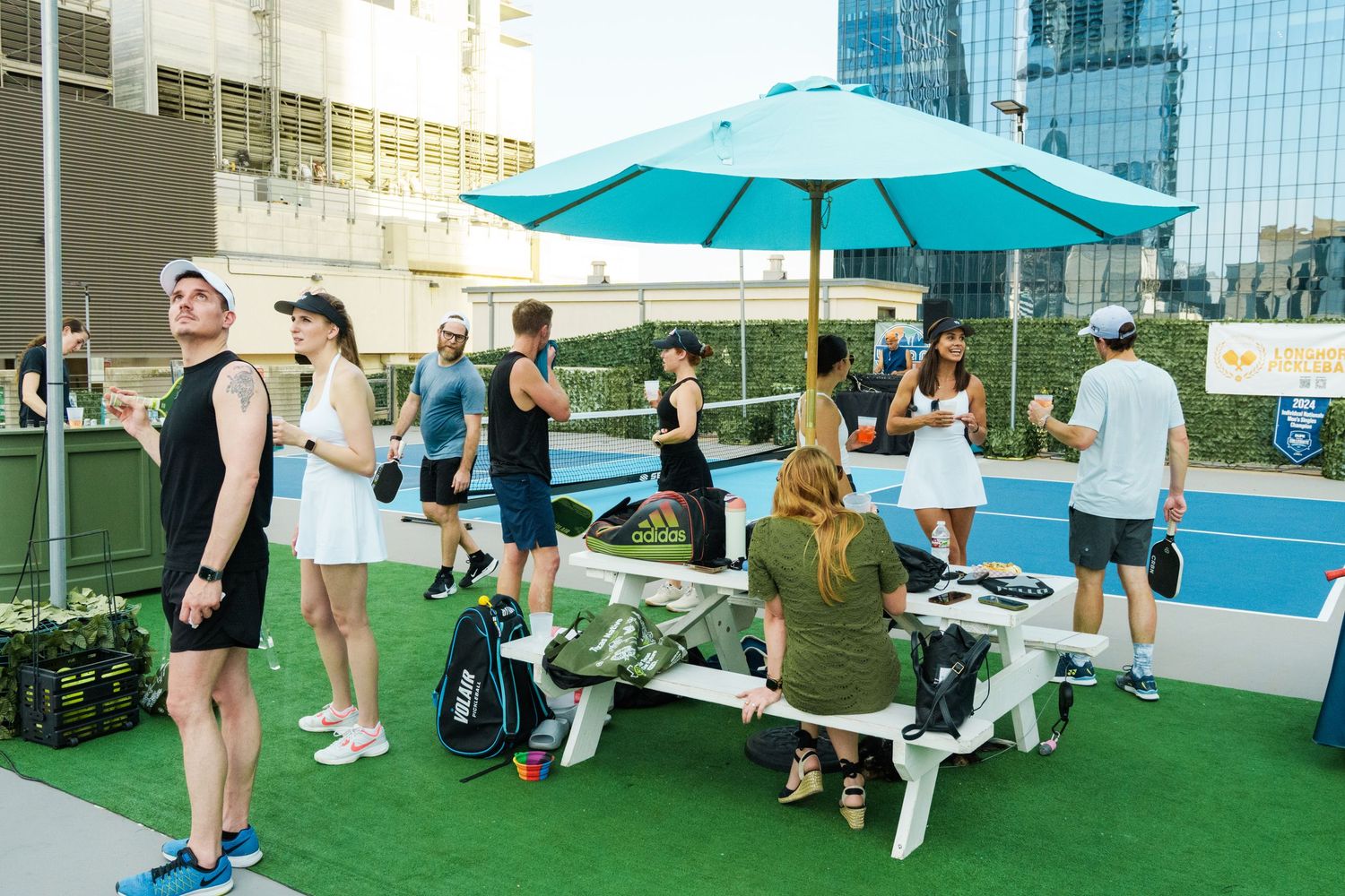 People socializing on a rooftop tennis court with a teal umbrella, picnic table, and city buildings in the background.