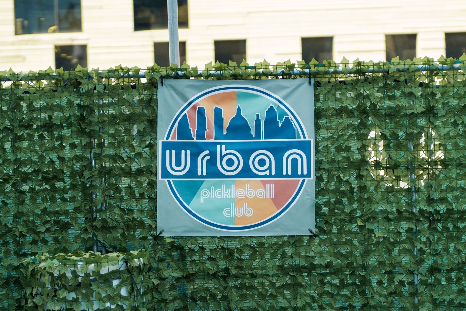 Urban Pickleball Club logo sign displayed on a green artificial hedge fence outdoors.