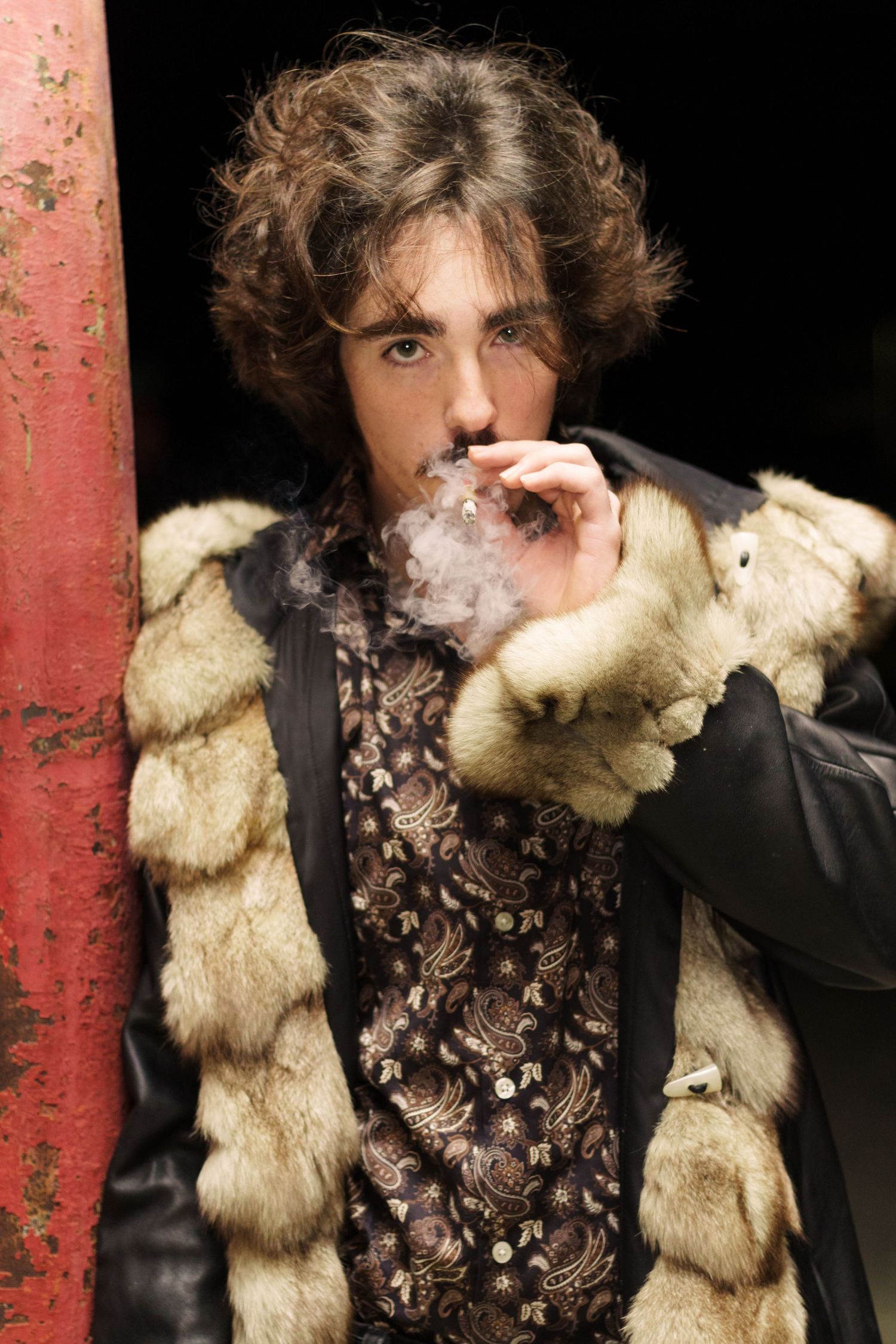 Mysterious man in fur-trimmed coat smoking against dark industrial background with red pillar.