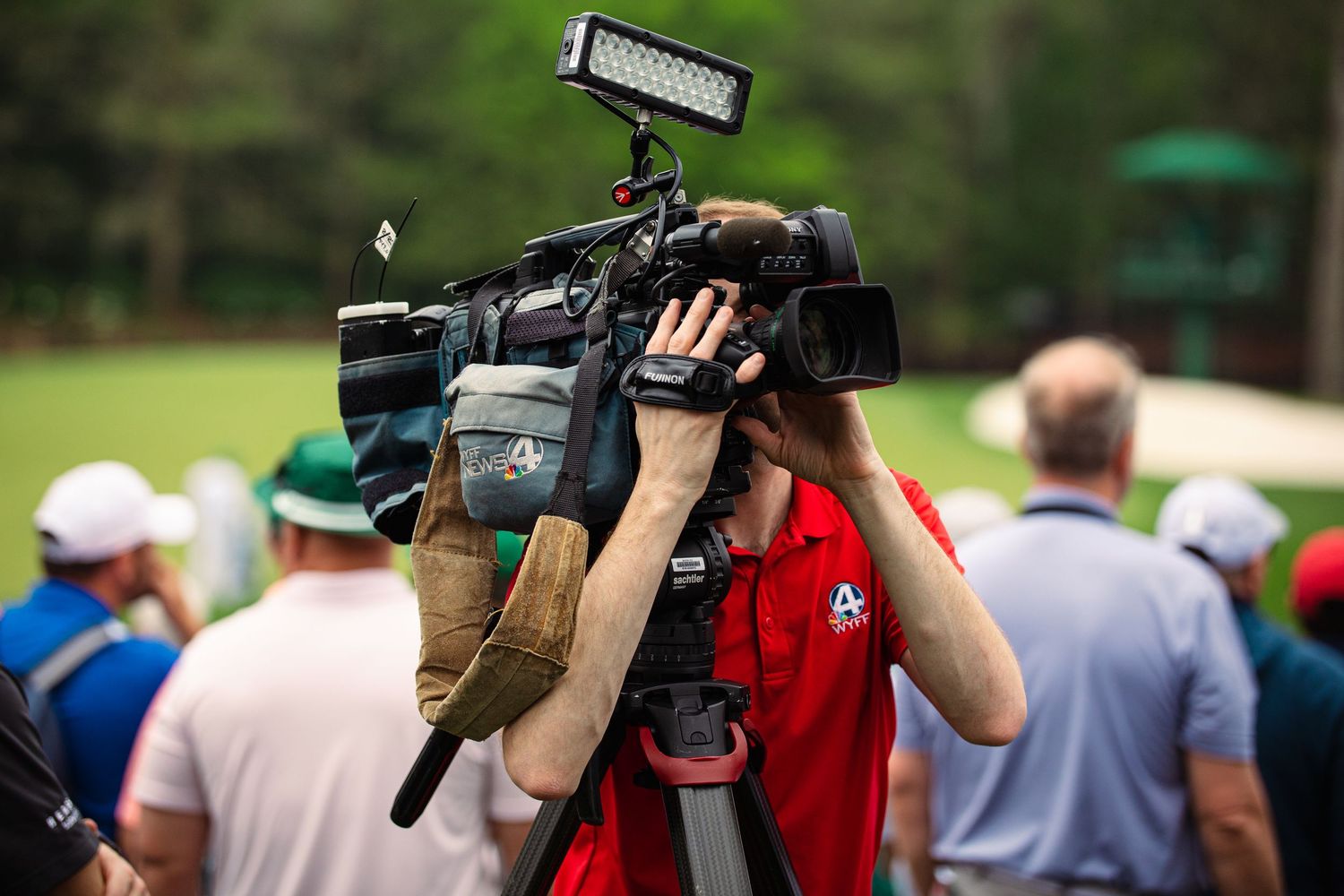 Cameraman in red shirt films golf tournament at Augusta National with professional video camera on tripod.