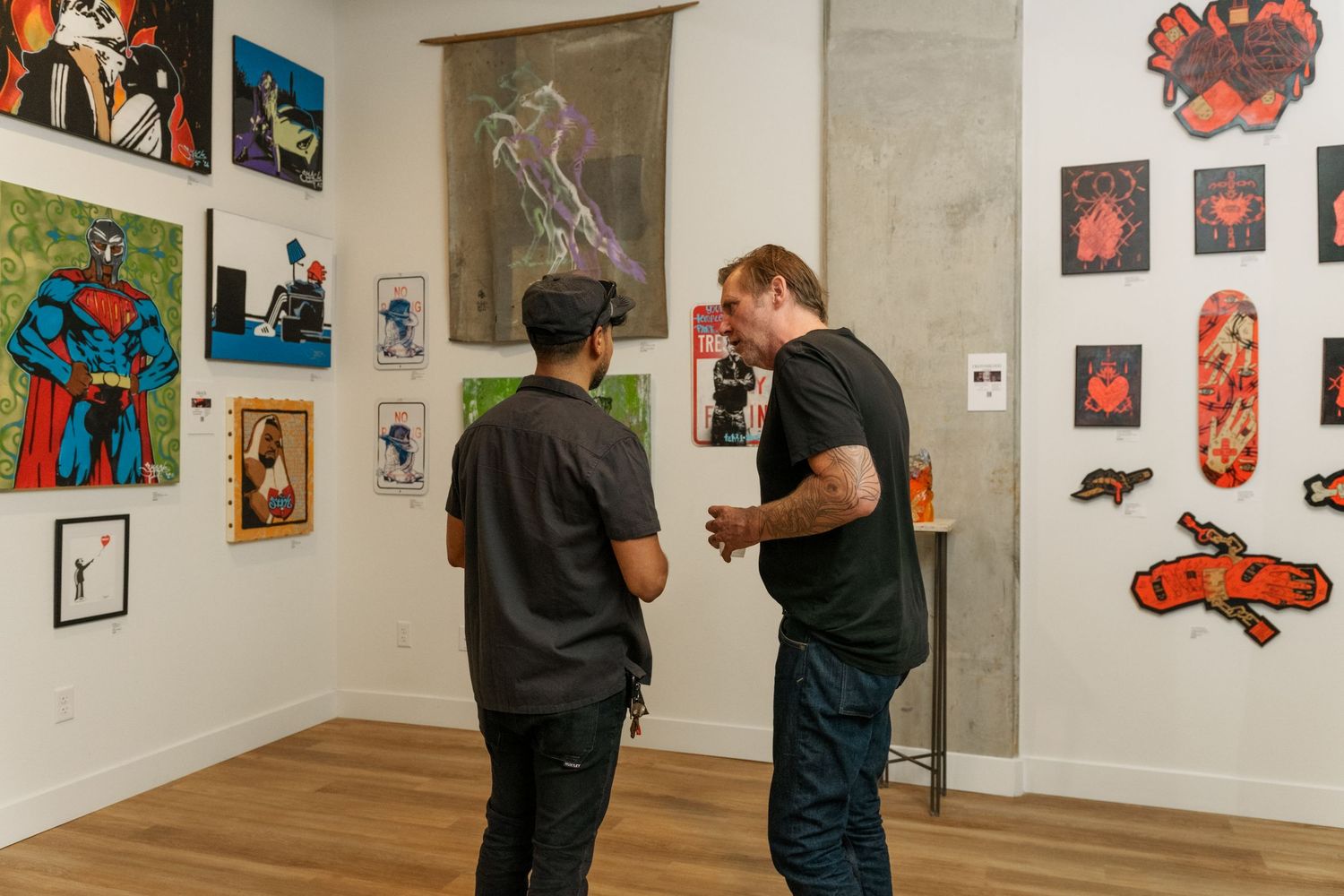 Two men converse in front of colorful superhero and pixel art at a gallery exhibition.