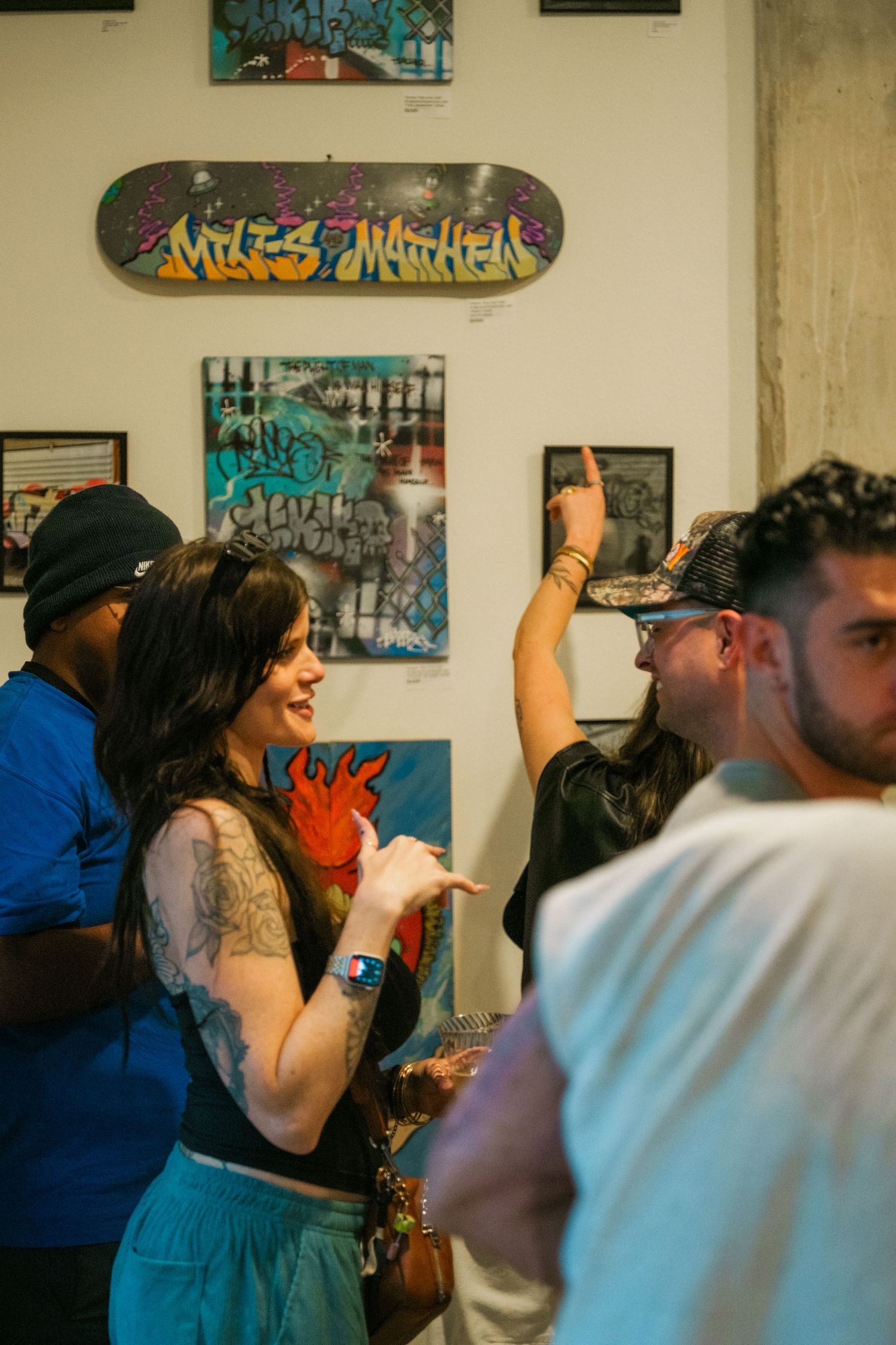 Tattooed woman laughs with guests at street art gallery opening featuring graffiti skateboard deck and urban paintings.
