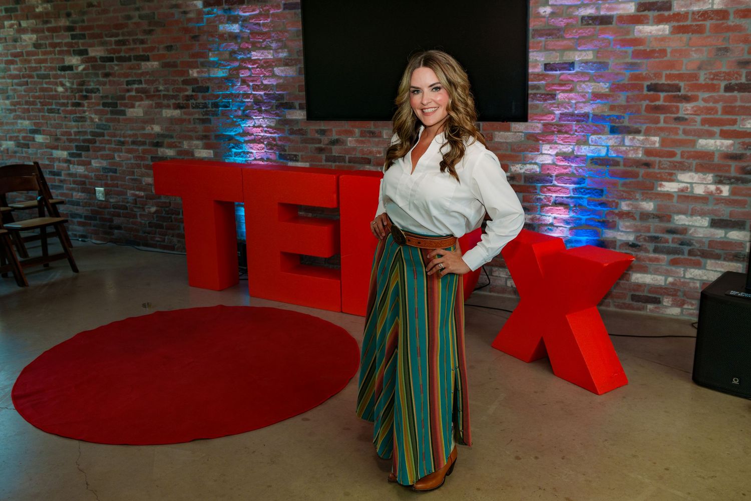 Hannah Hembree Bell in colorful striped skirt poses on red circle stage at TEDx event with brick wall backdrop. Round Rock Texas.
