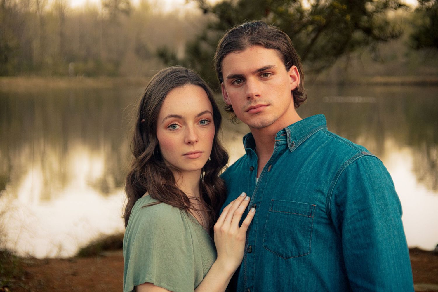 Couple posing seriously by a peaceful lake, woman in sage dress leaning against man in teal shirt.