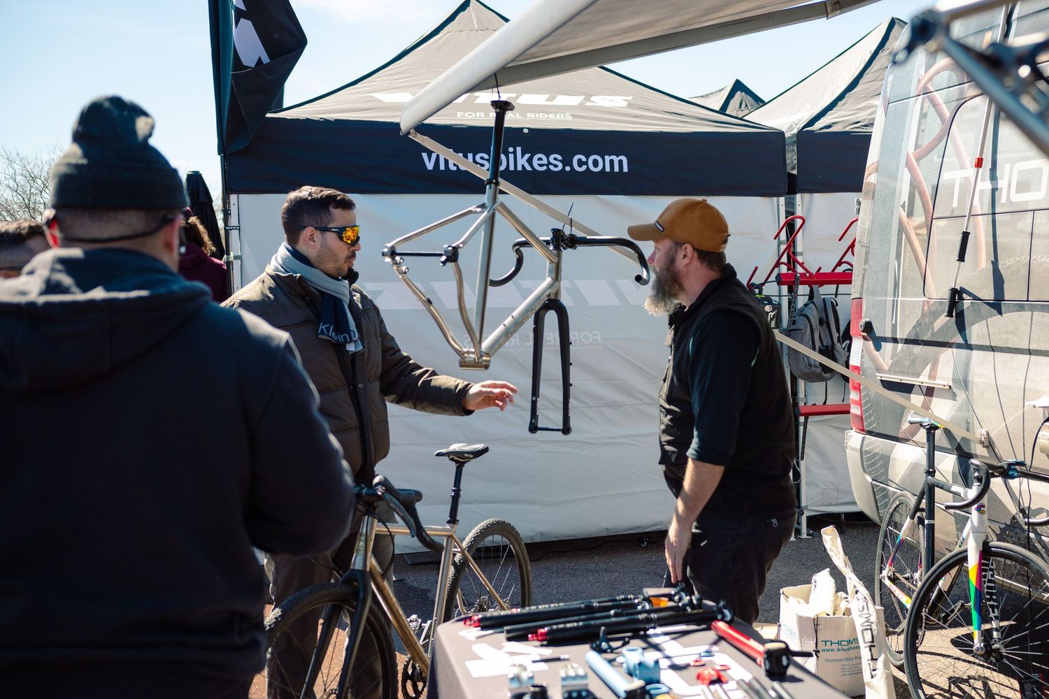 Cyclist speaking with a vendor at a Vitus Bikes outdoor expo booth displaying bicycle frames and components.