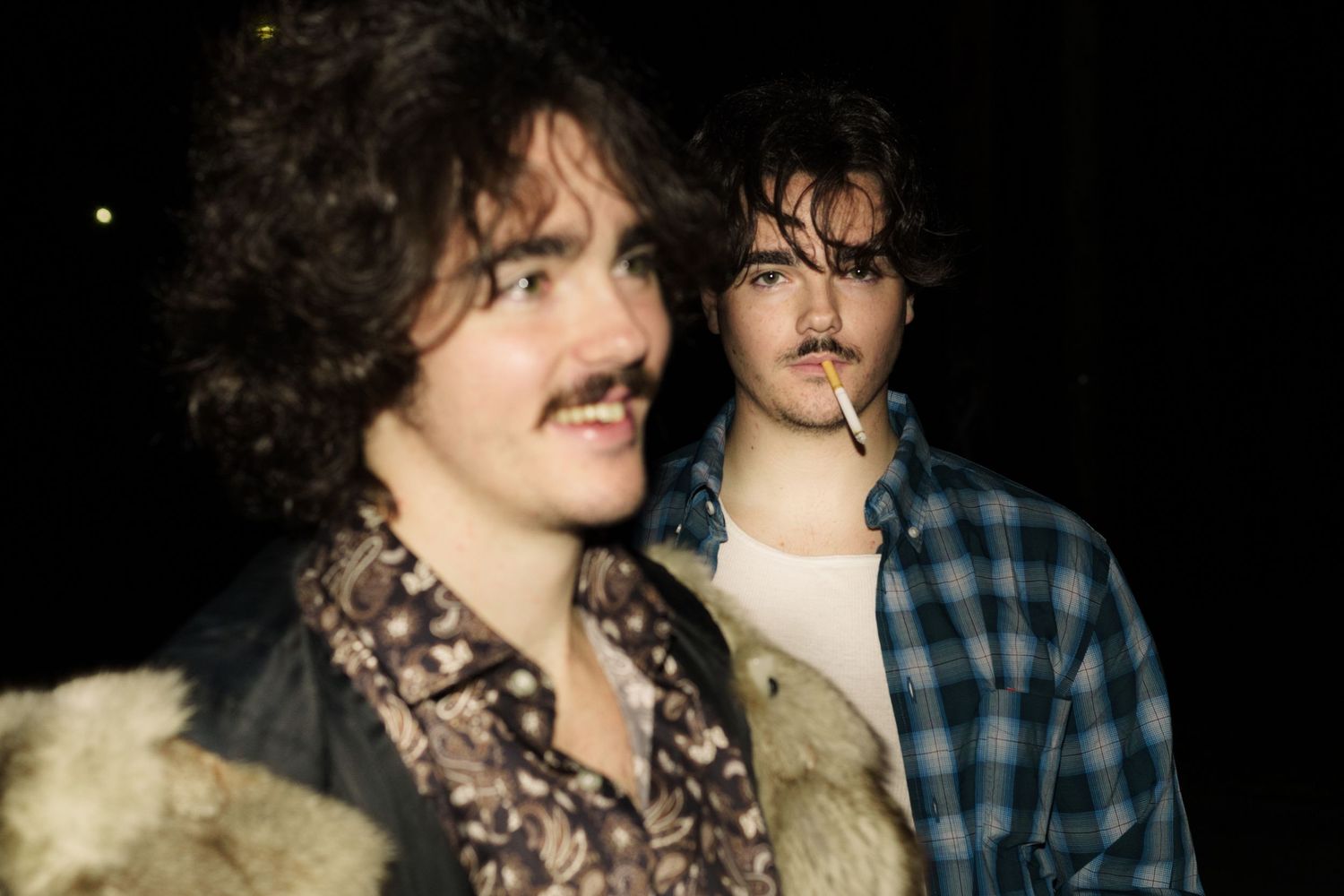 Two young men with curly hair stand together in dark setting.