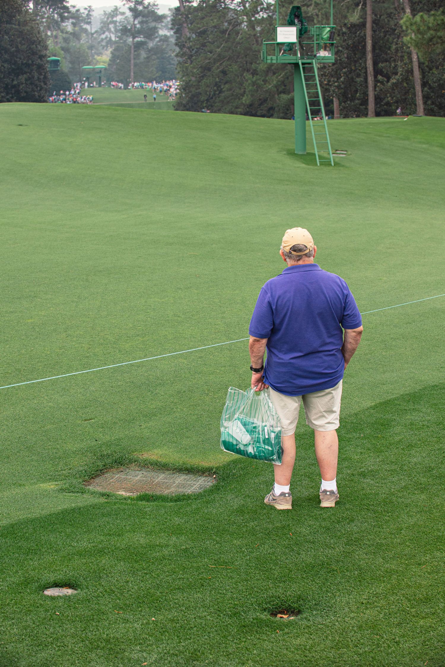 A groundskeeper in a blue polo shirt carries a bag of sand to repair a divot on a lush green golf course.