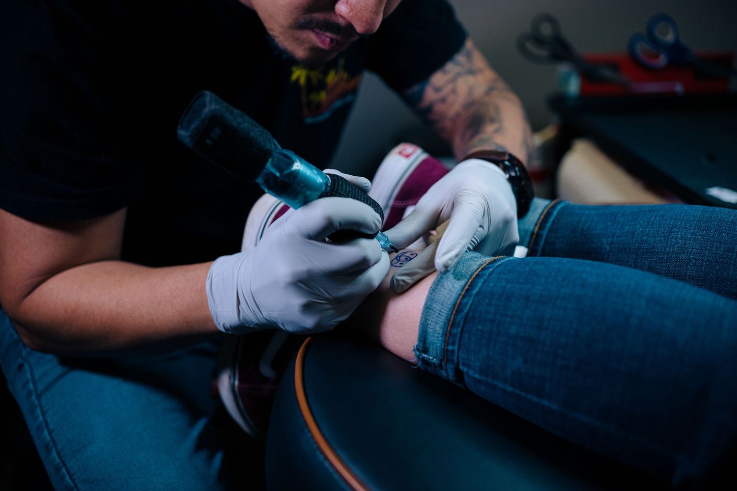 Tattoo artist applying ink to a client's leg using a tattoo machine while wearing white gloves.